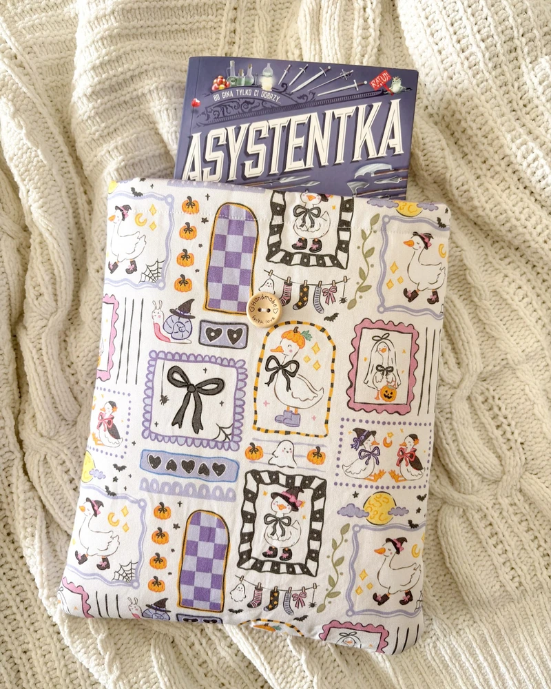 Booksleeve halloweenowe gąski, ze wzorem w gąski, duszki i dynie, z wystającą książką 'Asystentka złoczyńcy', na kocu