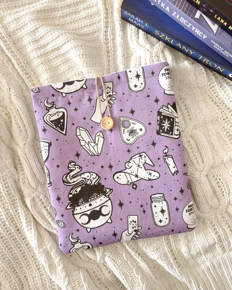 Booksleeve Kocioł czarownic w fiolecie ze wzorem w eliksiry, kociołki i gwiazdki, ułożony na kocu obok stosu książek