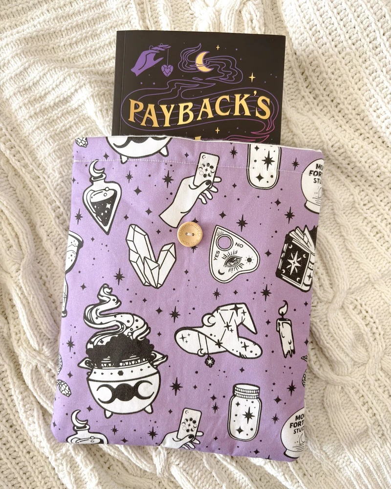 Booksleeve Kocioł czarownic w fiolecie ze wzorem w eliksiry, kociołki i gwiazdki, obok książki 'Payback's a witch', na kocu