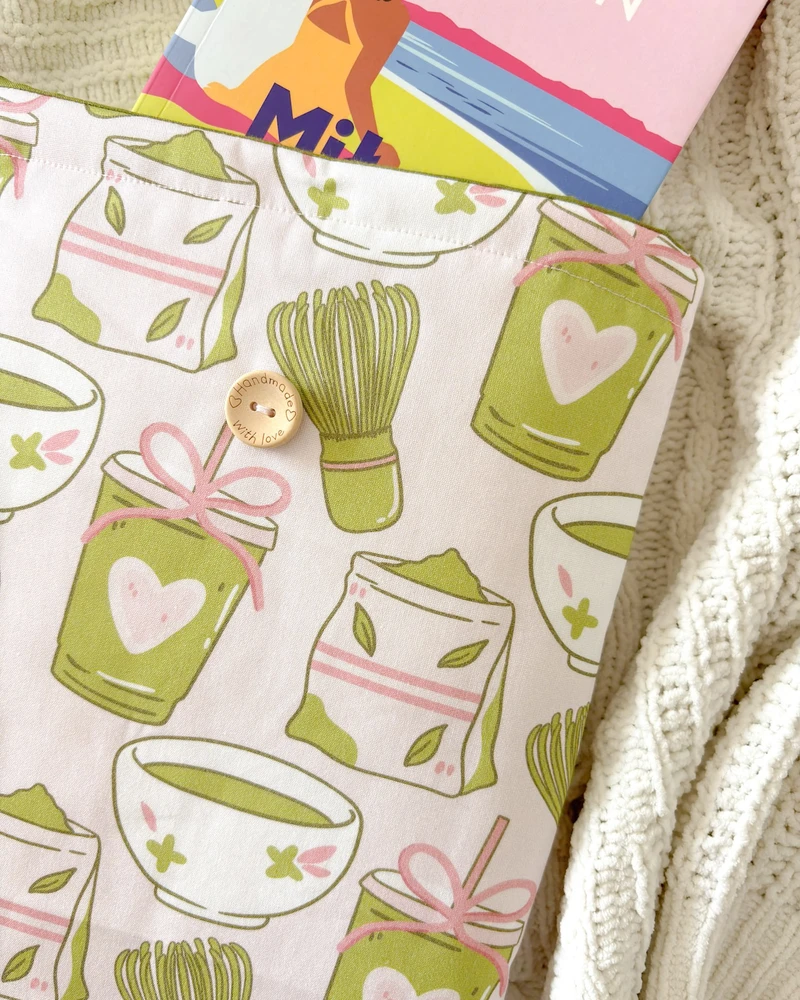 Booksleeve z wzorem w zielone kubeczki matcha, z wystającą książką, na kocu