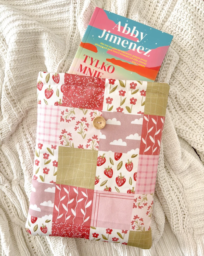 Booksleeve Patchwork w różowym patchworkowym wzorze z truskawkami i kwiatkami, z książką Abby Jimenez, na kocu