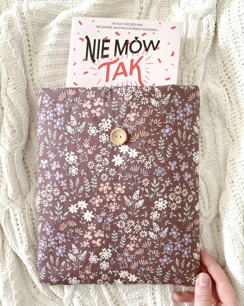 Booksleeve w brązowo-bordowym kolorze z drobnym wzorem w kwiaty, z wystającą książką 'Nie mów tak', na kocu