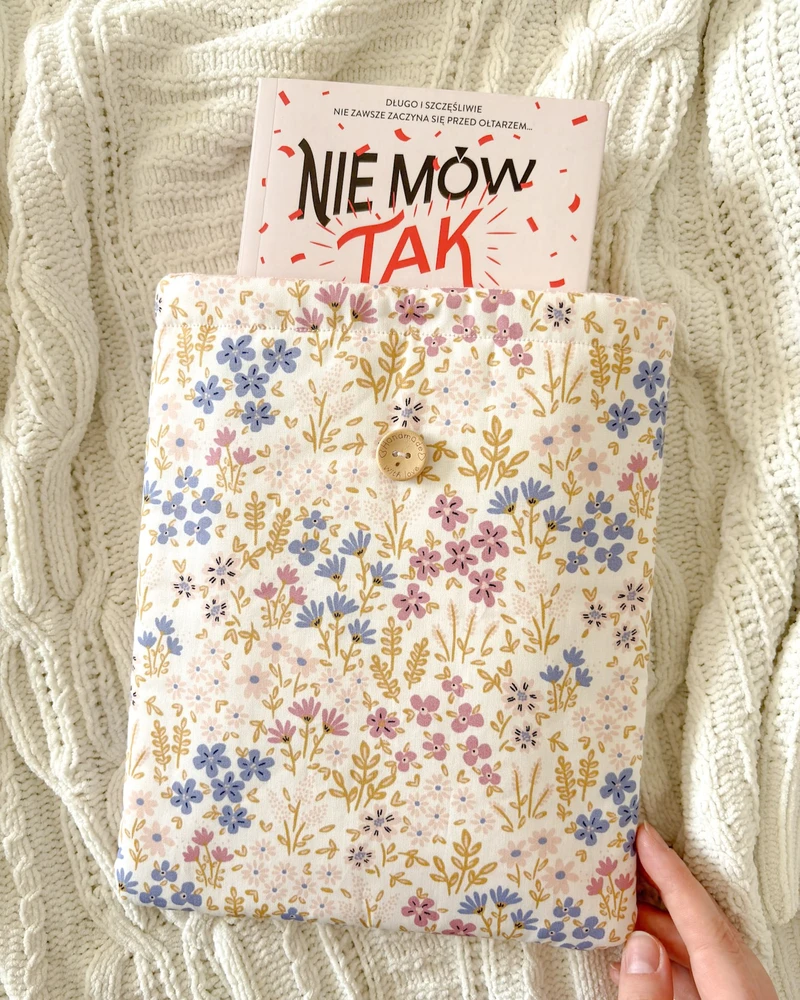 Booksleeve w kremowym kolorze z wzorem w kwiaty polne, z wystającą książką 'Nie mów tak', na kocu