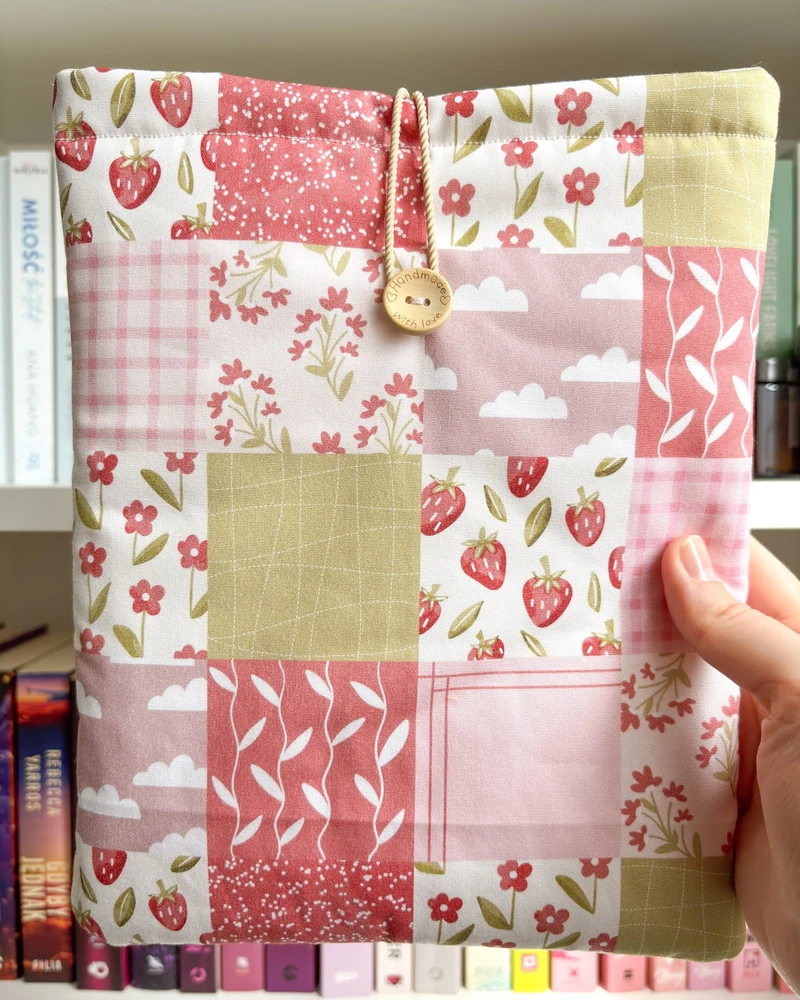 Etui na książkę Patchwork w różowym patchworkowym wzorze z truskawkami, trzymane w dłoni na tle regału z książkami