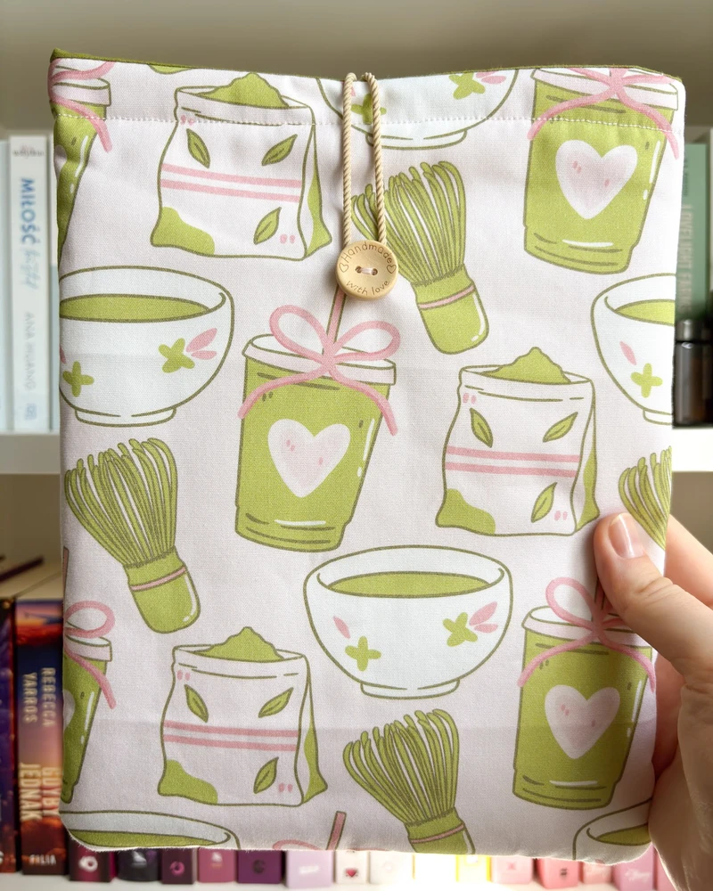 Etui na książkę w jasnoróżowym kolorze z wzorem w zielone kubeczki matcha, na tle regału z książkami