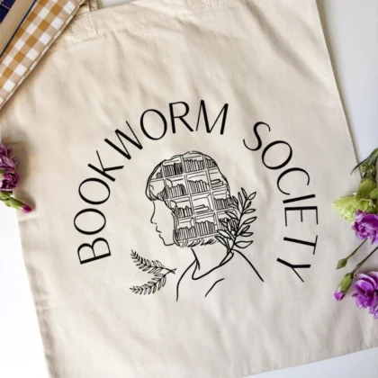 Torba bawełniana Bookworm Society z grafiką osoby, ułożona na stole wśród kwiatów
