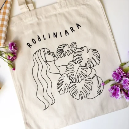 Torba bawełniana Rośliniara z grafiką liścia monstery, ułożona na stole wśród kwiatów