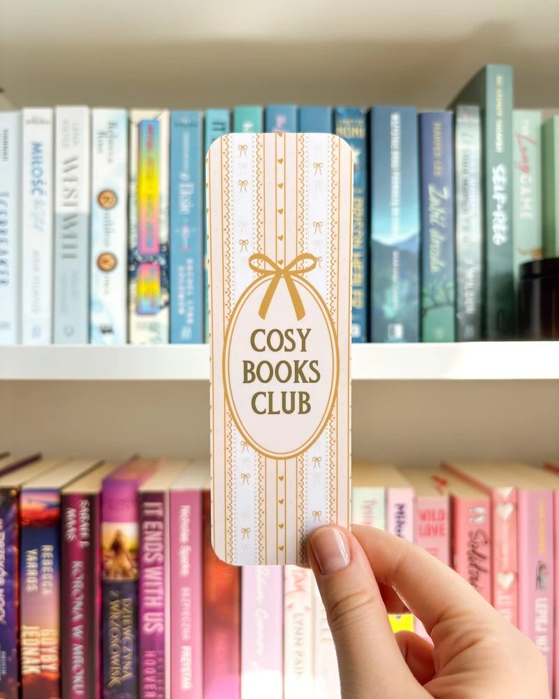 Zakładka w drobny żółty wzór i z ramką z napisem 'COSY BOOKS CLUB', trzymana w dłoni na tle regału z książkami