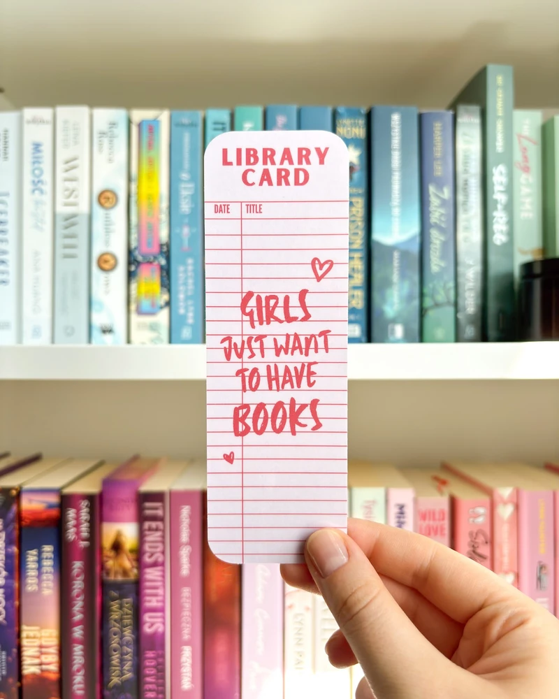 Zakładka w stylu karty bibliotecznej z napisem "girls just want to have books" w różowym kolorze, trzymana w dłoni na tle regału z książkami