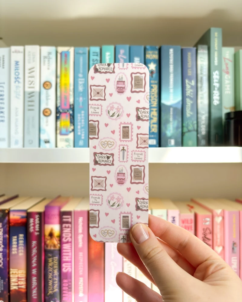 Zakładka w jasnoróżówym kolorze ze znaczkami book tropes, trzymana w dłoni na tle regału z książkami