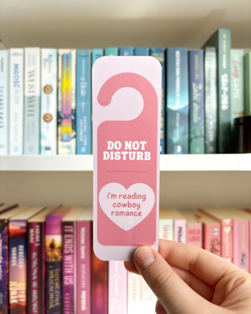 Zakładka w kształcie zawieszki 'DO NOT DISTURB, i'm reading cowboy romance' w różowym kolorze, trzymana w dłoni na tle regału z książkami