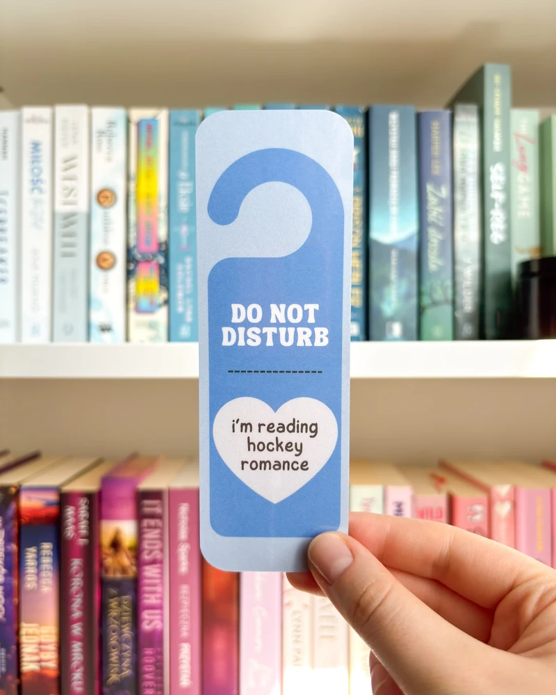 Zakładka w kształcie zawieszki 'DO NOT DISTURB, i'm reading hockey romance' w niebieskim kolorze, trzymana w dłoni na tle regału z książkami