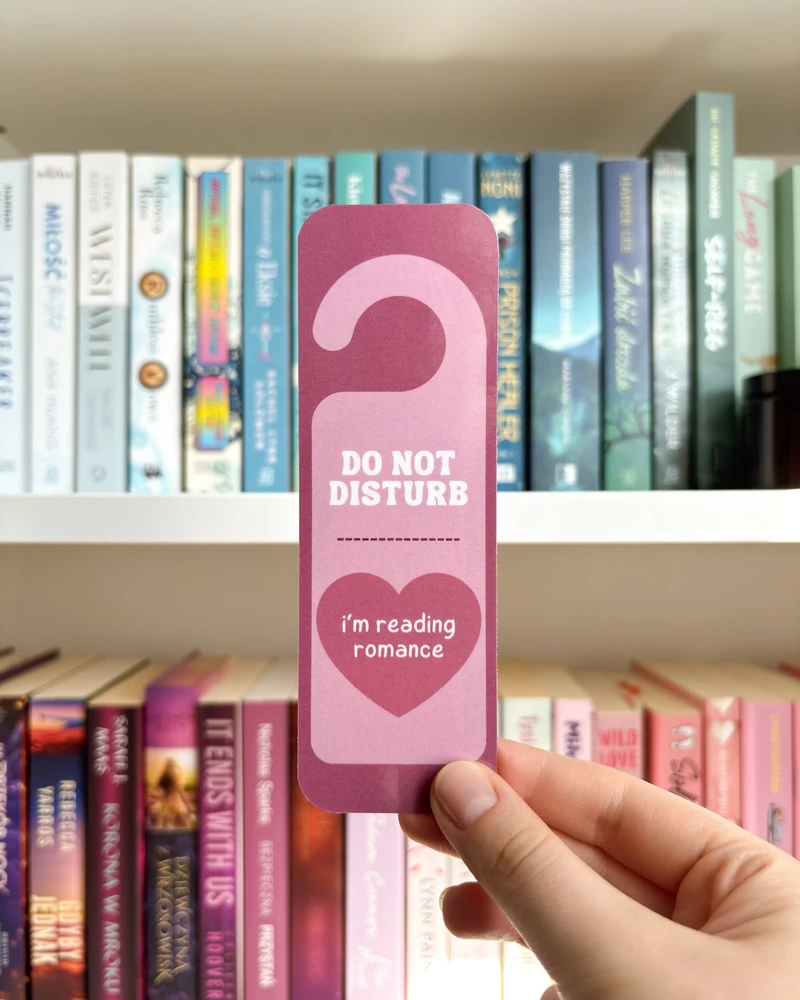 Zakładka w kształcie zawieszki 'DO NOT DISTURB, I'm reading romance' w różowym kolorze, trzymana w dłoni na tle regału z książkami