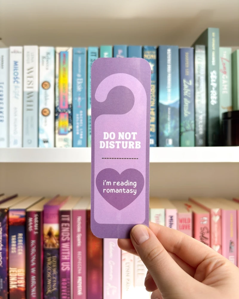 Zakładka w kształcie zawieszki 'DO NOT DISTURB, I'm reading romantasy' w jasnofioletowym kolorze, trzymana w dłoni na tle regału z książkami