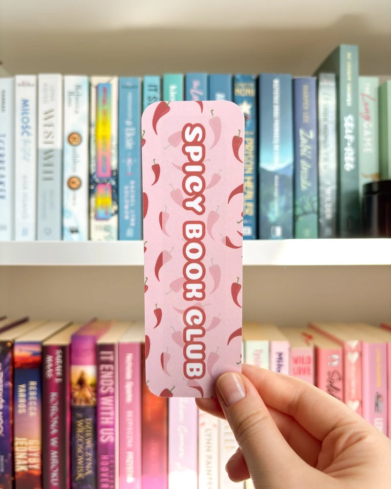 Zakładka w różowym kolorze z wzorem w papryczki chili i napisem 'SPICY BOOK CLUB', trzymana w dłoni na tle regału z książkami